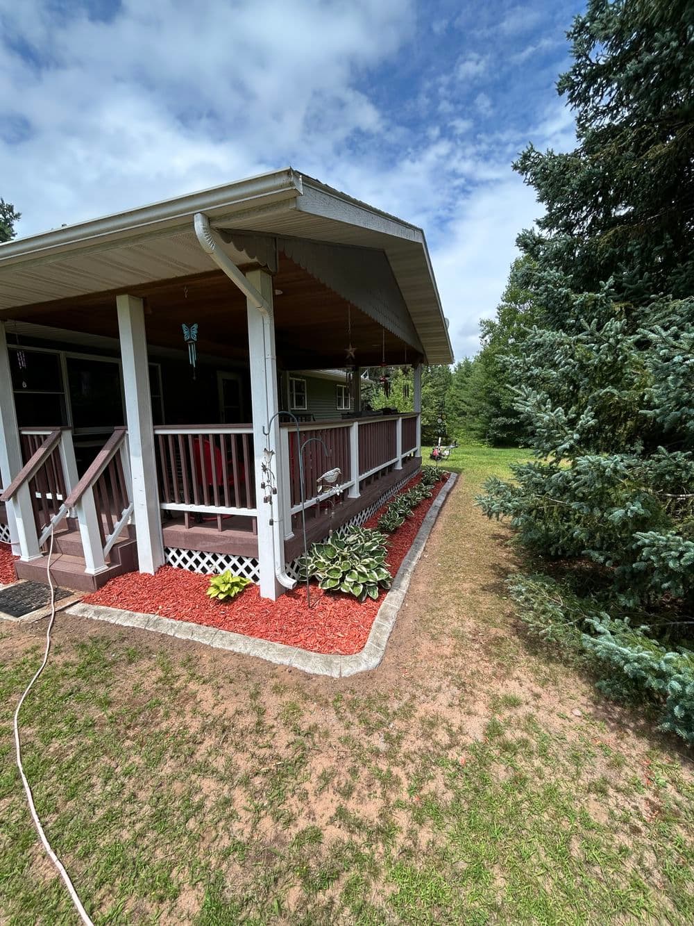 Charming cottage porch with flower beds and green trees under a partly cloudy sky.