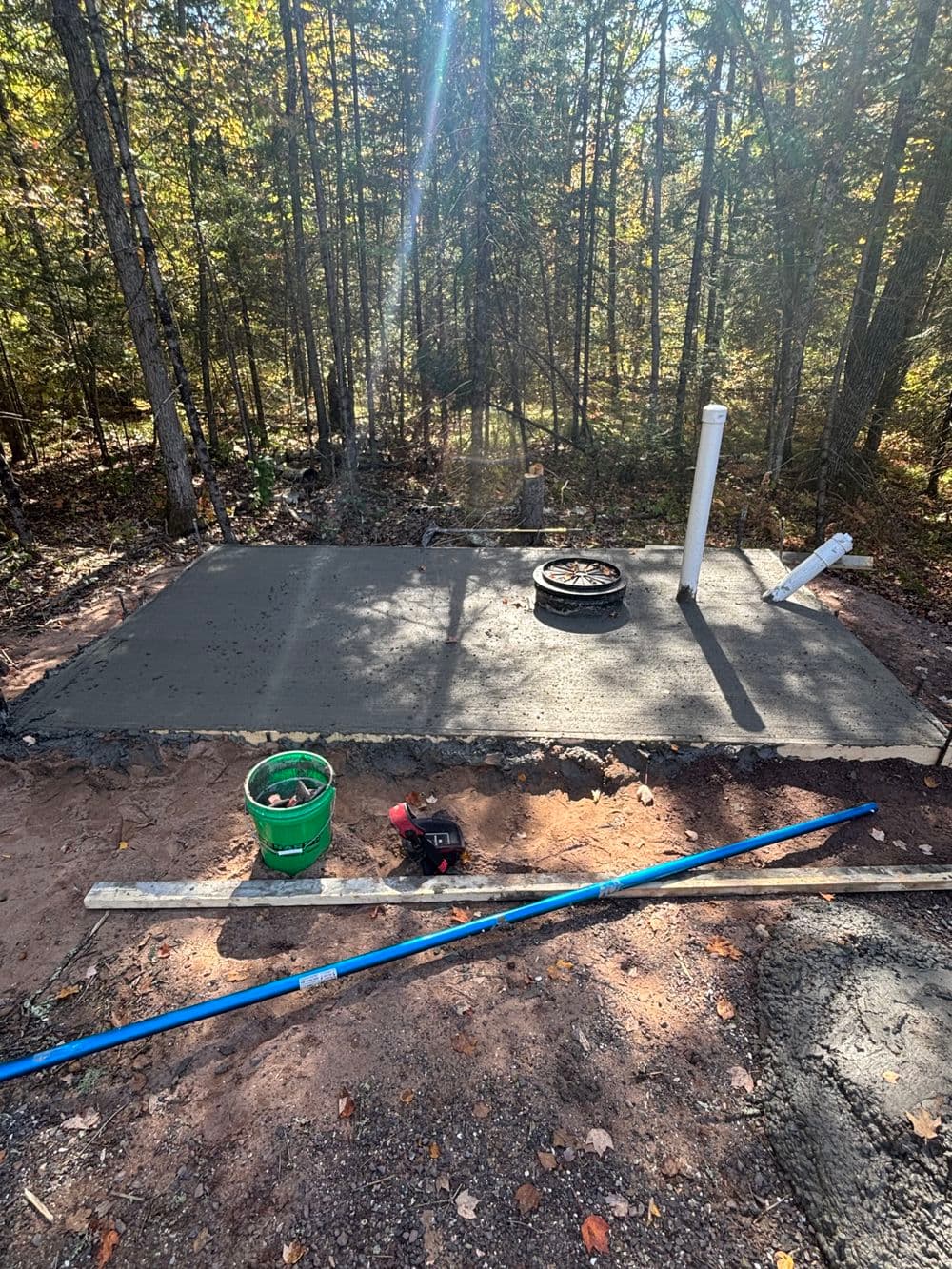 Concrete slab curing in wooded area, construction tools and materials nearby.