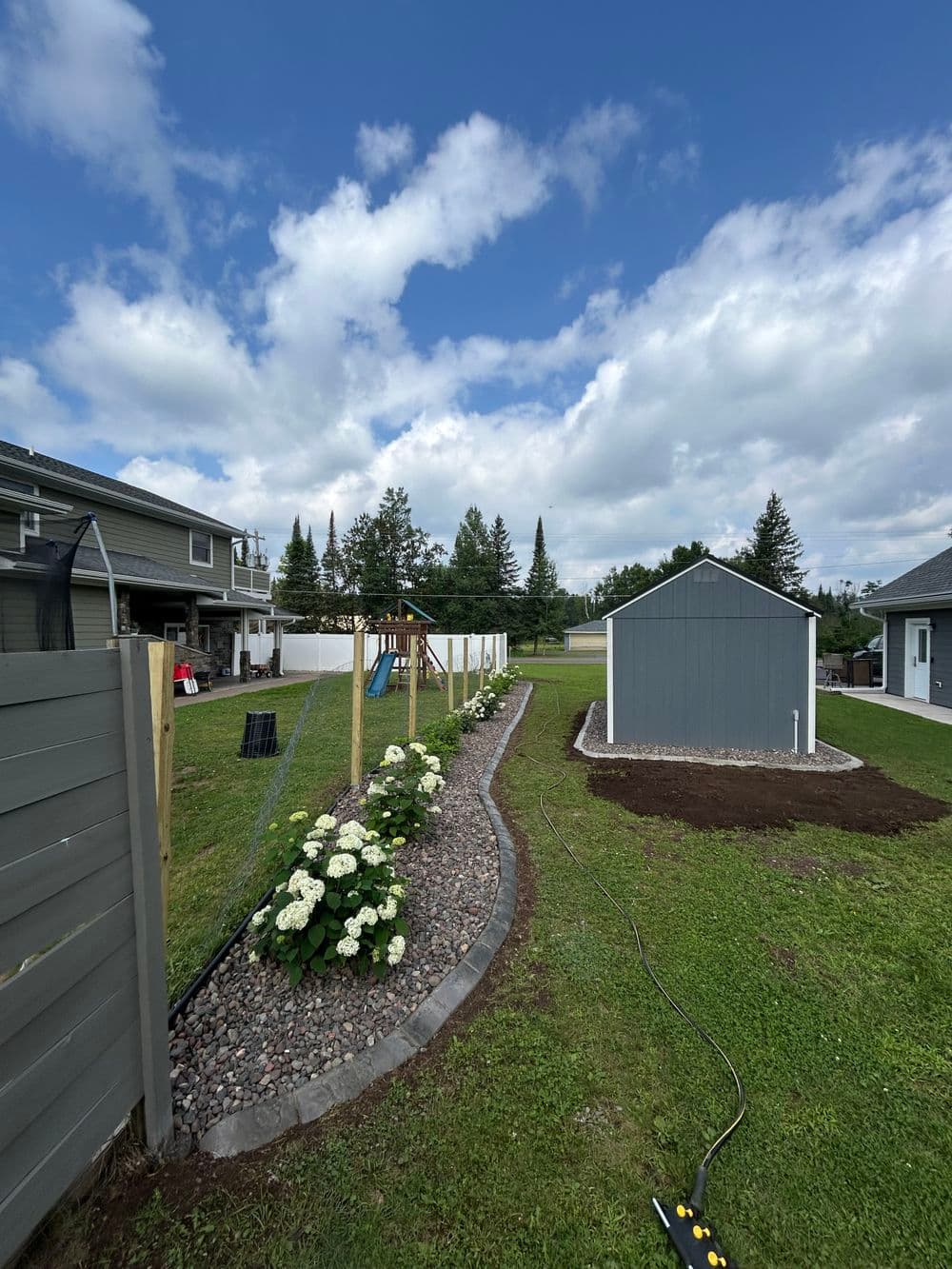 Lush backyard landscape with flowers, shed, and playset under a blue sky with clouds.