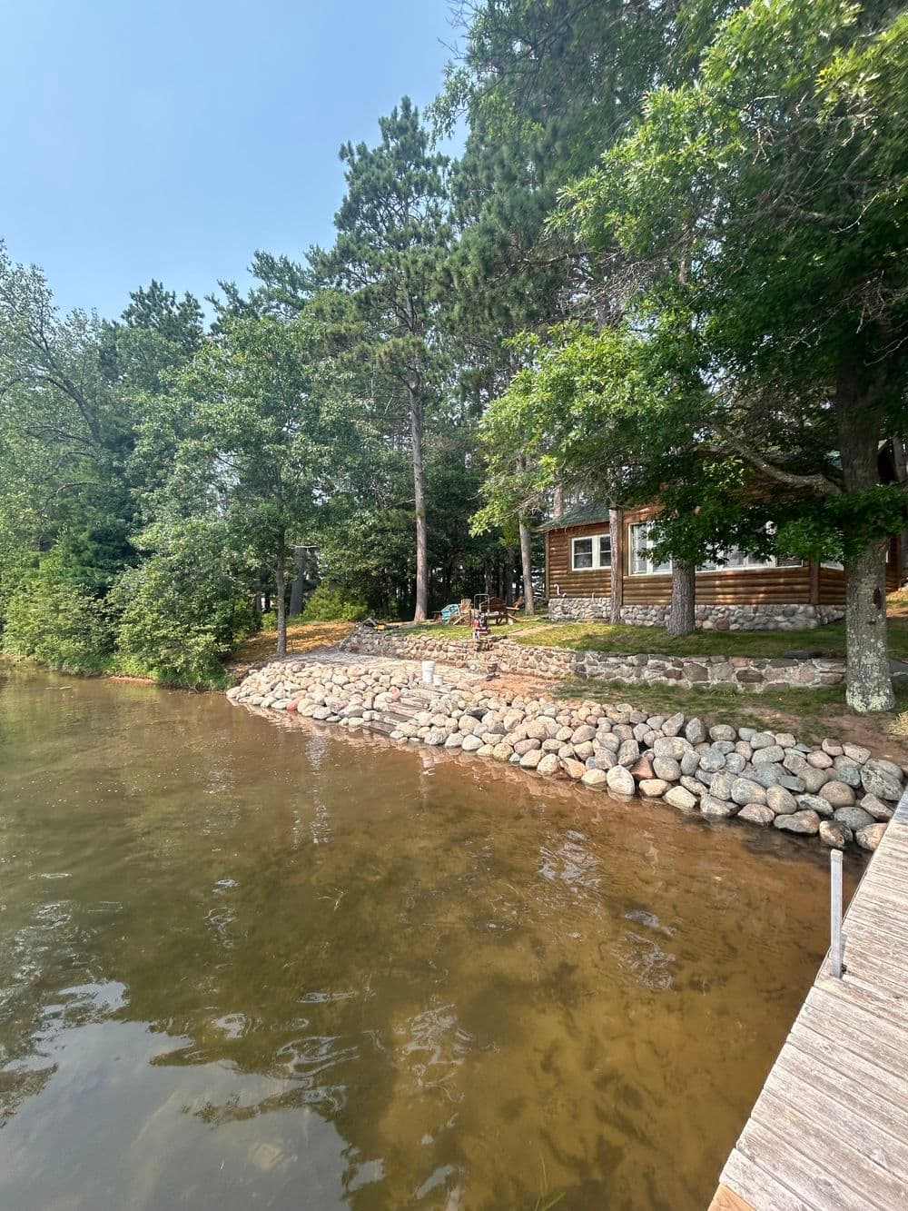 Lakeside cabin with rocky shoreline and lush greenery on a sunny day.
