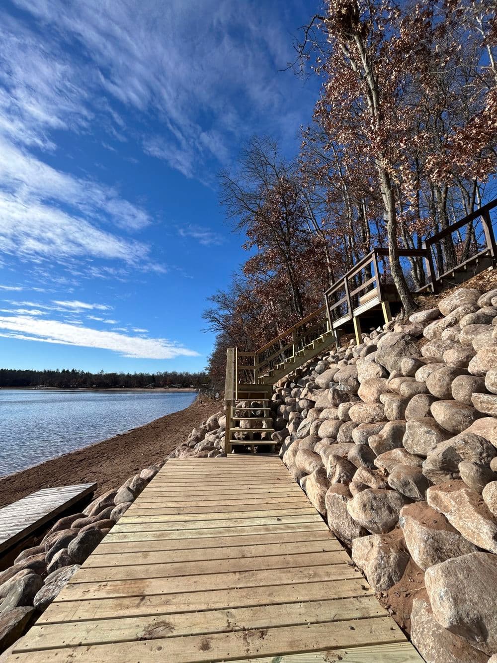 Wooden pathway leading to a serene lakeside with rocky shore and trees under a blue sky.