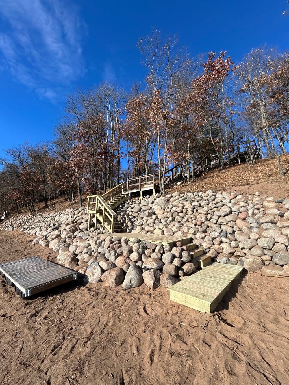Wooden stairs lead up a rocky shoreline under a blue sky with autumn trees.