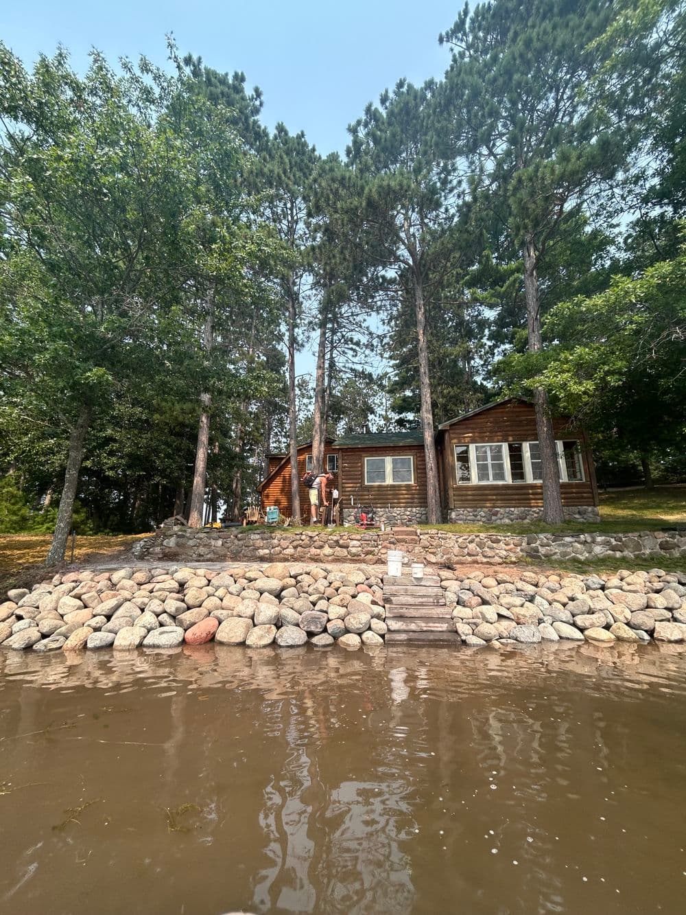 Lakeside cabin surrounded by trees with people enjoying outdoor activities by the water.