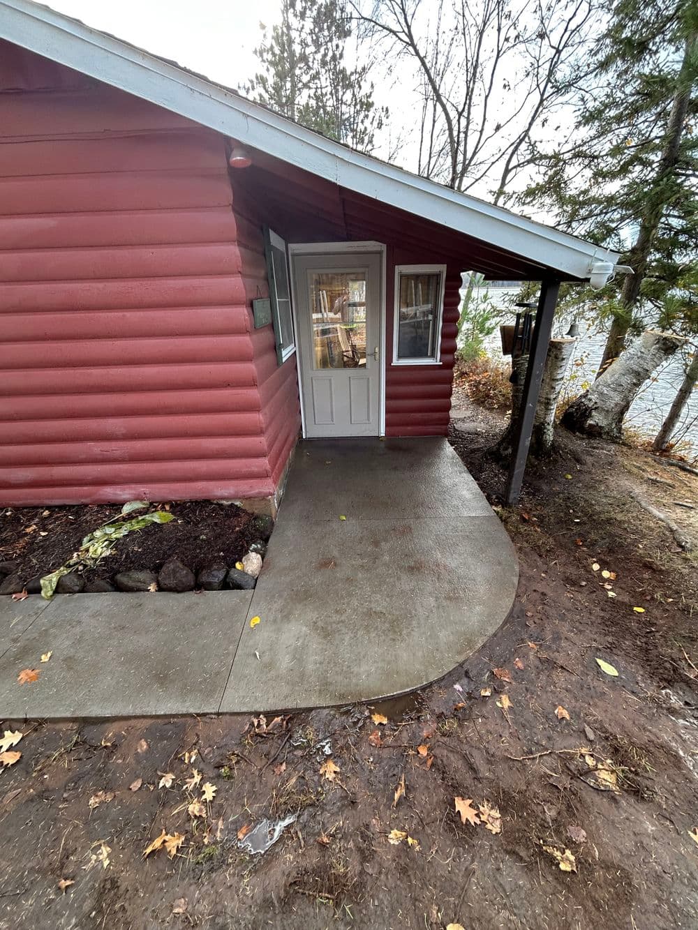 Red log cabin exterior with a concrete path and door, surrounded by trees and autumn leaves.